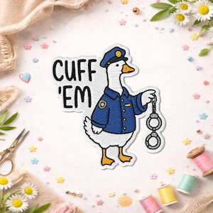 May include: Embroidered patch featuring a goose in a police uniform holding handcuffs. The goose wears a blue uniform and cap, with the words "CUFF 'EM" above. The patch is white with black and blue stitching.