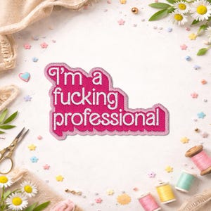 Peut inclure: Un écusson brodé rose avec le texte "I'm a fucking professional" en blanc. Le patch a un bord festonné. L'image comprend des éléments décoratifs tels que des fleurs, des bobines de fil et des ornements en forme d'étoiles.