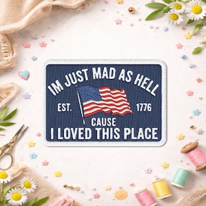 May include: A navy blue rectangular patch with white text that reads "IM JUST MAD AS HELL CAUSE I LOVED THIS PLACE." An American flag graphic is in the center. The patch also includes the text "EST. 1776."