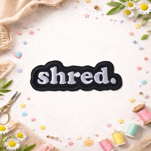 May include: A black embroidered patch with the word "shred." in white, stylized lettering. The patch has a black border and is surrounded by craft supplies, including spools of thread, scissors, and flowers.