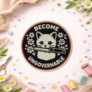 May include: Embroidered patch with a black background and a white cat holding a knife. The patch has the words "BECOME UNGOVERNABLE" in white and red. The patch is surrounded by white flowers and a red border.