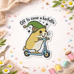 May include: An embroidered patch featuring a tan hamster wearing a green pointed hat with a yellow pom-pom, riding a light blue scooter. The text above the hamster reads, Off to cause a kerfuffle, accompanied by small black star icons.