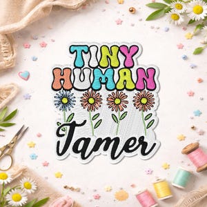 May include: Embroidered patch with the words "TINY HUMAN TAMER" in colourful bubble letters and a floral design. The patch features a white background with a stitched border and is surrounded by sewing supplies and decorative elements.