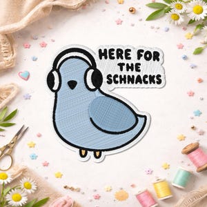 May include: An embroidered patch featuring a light blue pigeon wearing black and white headphones. To the right of the bird, a white speech bubble contains the black embroidered text "HERE FOR THE SCHNACKS" in a bold, sans-serif font.