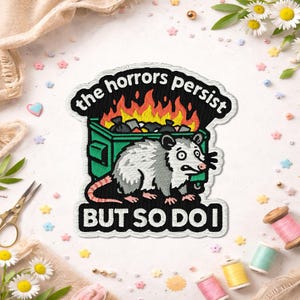 Peut inclure: Écusson brodé avec un opossum de dessin animé devant une poubelle en feu. Le texte "the horrors persist BUT SO DO I" est affiché. Le patch a une bordure noire et est posé sur un fond blanc avec des éléments décoratifs.