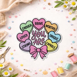 May include: Embroidered patch featuring the words "You Are" surrounded by heart-shaped candies with affirmations such as "Brave" and "Worthy." A pink bow is at the bottom. Daisies and sewing supplies surround the patch.