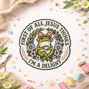 May include: A circular embroidered patch featuring a green frog wearing a pink bow, holding a small cup, and surrounded by a floral wreath. The text reads: FIRST OF ALL JESUS THINKS I'M A DELIGHT. The design is stitched with white, green, and pink thread.
