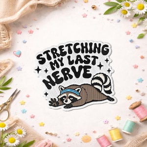 May include: Embroidered patch featuring a raccoon stretching, with the text "Stretching My Last Nerve" in black and white. The raccoon is brown, with a blue headband and a striped tail. The patch is surrounded by small, colorful embellishments.
