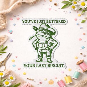 May include: A green embroidered patch featuring a frog wearing a cowboy hat and overalls, with the text "YOU'VE JUST BUTTERED YOUR LAST BISCUIT." The patch is surrounded by decorative elements like flowers and spools of thread.