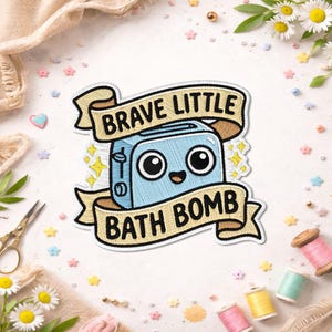 May include: Embroidered patch featuring a light blue toaster with large eyes and a smiling mouth. The patch reads "BRAVE LITTLE BATH BOMB" on a banner. The design includes yellow starbursts and is surrounded by craft supplies.