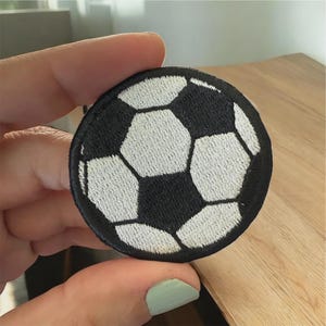 May include: A circular embroidered patch featuring a classic black and white soccer ball pattern. The design consists of alternating hexagonal and pentagonal shapes stitched with white and black thread, finished with a solid black stitched border.