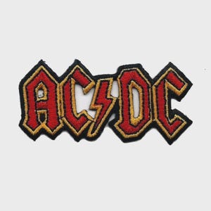 May include: Embroidered patch featuring the band name "AC/DC" in red, outlined in gold, with a black border. The design includes a lightning bolt in the center, a classic rock and roll emblem.
