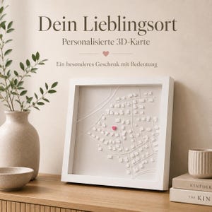 Könnte beinhalten: Eine quadratische, weiß gerahmte 3D-Karte mit erhabenen weißen Gebäudeblöcken und Straßenlinien auf hellem Untergrund, wobei ein kleines rosa Herz eine der Strukturen markiert. Der Text darüber lautet "Dein Lieblingsort", "Personalisierte 3D-Karte" und "Ein besonderes Geschenk mit Bedeutung".