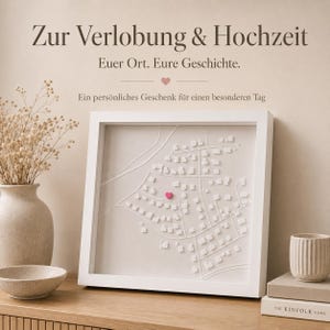 Könnte beinhalten: Ein quadratisches, weiß gerahmtes Wandbild mit einer 3D-Reliefkarte eines Stadtplans auf hellem Untergrund. Ein kleines rosa Herz ist auf einem der Gebäude platziert. Der Text darüber lautet: Zur Verlobung & Hochzeit, Euer Ort. Eure Geschichte.