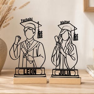 Könnte beinhalten: Zwei Holzfiguren zum Thema Abschluss. Eine zeigt einen Absolventen mit dem Text "Fachabi" und dem Namen "Leon". Die andere zeigt einen Absolventen mit dem Text "Abitur" und dem Namen "Julia". Beide Figuren tragen das Jahr "2026" auf ihren Abschlusskappen.