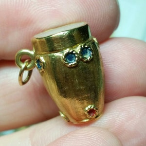 May include: A gold-colored charm in the shape of a bongo drum with blue and red enamel accents. The charm has a loop for attaching to a necklace or bracelet.