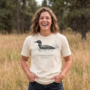 May include: A cream-colored t-shirt featuring a detailed black and white illustration of a Common Loon swimming in water. The text "Common Loon Gavia immer" is printed below the illustration. The shirt is worn by a person in a natural outdoor setting.