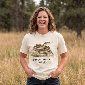 May include: A cream-colored short-sleeve t-shirt featuring a graphic illustration of a garter snake coiled on a rock. The text "garter snake sunbath" is printed in a dark, rounded font directly below the snake illustration on the front.