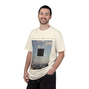 May include: Off-white t-shirt with a graphic of a black square hovering over a city skyline against a cloudy sky. The text "Government to release alien files." is printed above the image. The shirt is a short-sleeved, casual design.