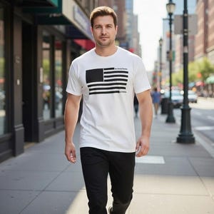 Peut inclure: Un t-shirt blanc avec un drapeau américain noir et blanc. Le t-shirt est à manches courtes et est porté avec un pantalon noir. Le texte "The United States of America" est au-dessus du drapeau.