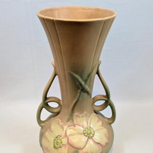 May include: A ceramic vase with a flared rim and two curved handles. The body features a matte finish in muted tan and green tones, decorated with embossed pale yellow flowers with pink edges and green centres, arranged in a symmetrical pattern.