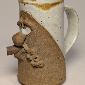 May include: A handmade ceramic mug with a whimsical face design. The mug features a white and brown color scheme, with a handle on the side. The face has a large nose, eyes, and a curly mustache. The mug is approximately 5 inches tall.