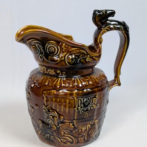 May include: A brown ceramic pitcher with a decorative handle and spout. The pitcher features embossed floral and figural designs, with a dark brown glaze. The handle is topped with a small animal figure.