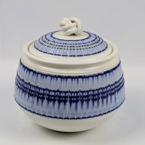 May include: A round ceramic sugar bowl with a lid featuring a knot-shaped handle. The bowl and lid are decorated with a repeating pattern of vertical blue lines and horizontal bands against a cream-colored background, creating a textured appearance.