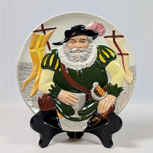 May include: A decorative ceramic plate featuring a detailed relief of a man in historical attire. The plate has a white background with a green, yellow, and brown color scheme. The plate is displayed on a black wooden stand.