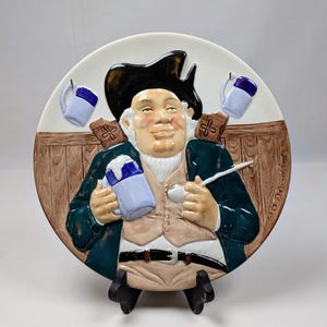 May include: A decorative ceramic plate featuring a jovial man in a black hat and green coat, holding a mug of beer and a pipe. The plate has a white background with two mugs and a wooden panel design. The plate is displayed on a black stand.
