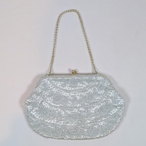 May include: A white handbag featuring an intricate silver floral embroidery pattern across the entire surface. The bag has a curved shape, a metallic silver chain strap, and a small gold-toned metal clasp closure at the top center.