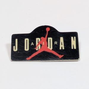 May include: A rectangular pin with a black background featuring the red Jumpman logo in the center. The text JORDAN is printed in large cream-colored letters across the background, with the word AIR written in smaller letters over the logo.
