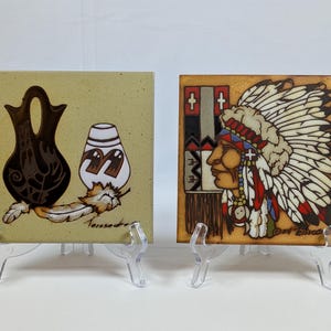 May include: Two square ceramic tiles displayed on clear plastic stands. The left tile features a dark brown pottery vessel, a white patterned jar, and a feather on a tan background. The right tile depicts a profile of a person in a feathered headdress.