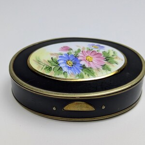 May include: An oval black compact case with a gold-toned metal rim and a central inset featuring a hand-painted floral design. The illustration displays pink and blue flowers with green leaves on a white background. A small gold latch is visible.