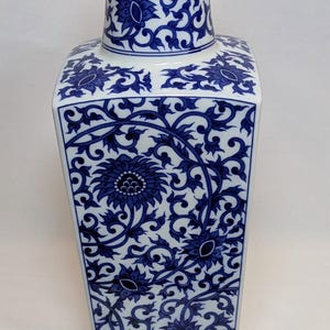 May include: A tall, square-shaped ceramic vase with a white background. The vase features a blue and white floral pattern. The pattern includes flowers and swirling vines. The vase has a narrow neck and a wide base. The vase is approximately 12 inches tall.