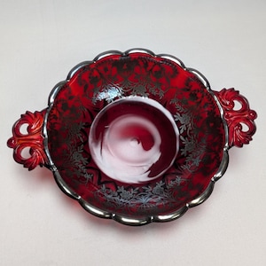 May include: A round, deep red glass bowl featuring two ornate, scrolled handles on opposite sides. The interior surface is decorated with a metallic silver floral pattern, and the scalloped rim is finished with a matching silver metallic edge.