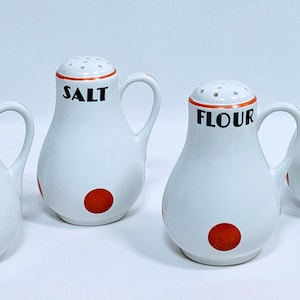 May include: Four white ceramic kitchen canisters with handles, labeled "PEPPER", "SALT", "FLOUR", and "SUGAR". Each has a red dot on the base and an orange band around the top. The canisters are designed for kitchen use.