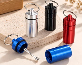 May include: A set of four cylindrical metal pill containers with keyrings, available in silver, black, red, and blue. One blue container is open, revealing a small attached metal spoon inside, while the others remain closed.