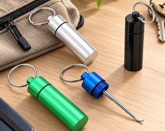 Könnte beinhalten: Ein Set aus vier zylindrischen Pillendosen-Schlüsselanhängern aus Metall in Silber, Schwarz, Grün und Blau. Jedes Teil hat einen Schraubdeckel mit Metallring. Das blaue Gehäuse ist aufgeschraubt und zeigt ein schmales, silbernes Metallwerkzeug.