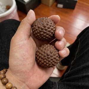 Puede incluir: Dos esculturas esféricas de madera marrón con superficies texturizadas, sostenidas en una mano. Las esculturas tienen un patrón detallado y con bultos, que se asemeja a una piña o una forma natural similar. Se ve una pulsera de cuentas de madera.