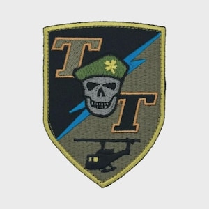 May include: Embroidered patch with a shield shape. The patch features a skull wearing a green beret, a helicopter, and the letter "T" in a military-style design. The patch has a black, olive green, and gold color scheme.