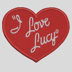 May include: Red heart-shaped patch with white embroidered text that reads "I Love Lucy". The patch has a stitched border and is likely for application on clothing or accessories. The design is a tribute to the classic television show.