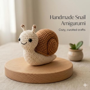 May include: A handmade crochet snail amigurumi in beige and brown. The snail has a smiling face and a spiral shell. The text "Handmade Snail Amigurumi" and "Cozy, curated crafts" are displayed above the snail.