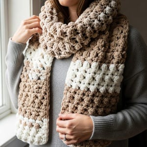 May include: A chunky, hand-crocheted scarf in a neutral color palette of cream, tan, and brown. The scarf features a textured stitch pattern and a wide, rectangular shape, perfect for wrapping around the neck. The scarf is a cozy winter accessory.