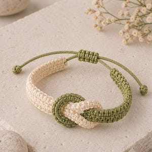 May include: A woven cord bracelet featuring a central knot design. The bracelet is constructed from alternating sections of cream and olive green thread, with a sliding knot closure mechanism that allows for adjustable sizing on the wrist.
