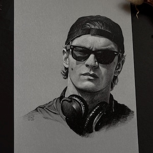 Può includere: Un disegno a grafite in bianco e nero su carta grigia che raffigura una persona con un berretto da baseball, occhiali da sole scuri e cuffie attorno al collo. Il ritratto presenta ombreggiature dettagliate e la firma dell'artista M.29 W.26.