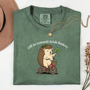 May include: Green t-shirt with the text "Off to commit Irish foolery" and a cartoon hedgehog riding a red tricycle, holding a four-leaf clover. A gold necklace with a star pendant is draped on the shirt.