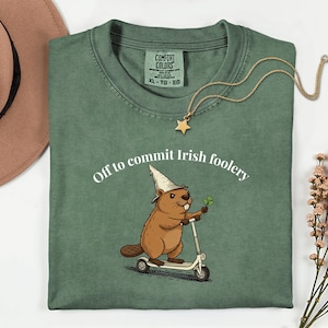 May include: Green t-shirt featuring the text "Off to commit Irish foolery" and a cartoon beaver wearing a wizard hat and holding a four-leaf clover while riding a scooter. A gold necklace with a star pendant is on the shirt.