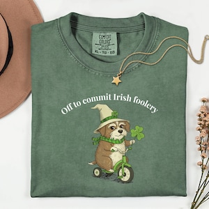 May include: Green t-shirt with the slogan "Off to commit Irish foolery." The shirt displays a cartoon dog in a leprechaun hat and scarf, riding a green tricycle and holding a four-leaf clover. A gold star pendant necklace is draped on the shirt.