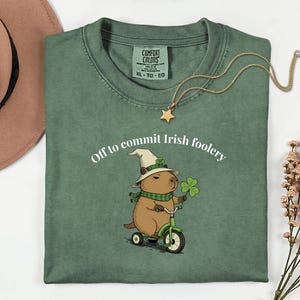 Peut inclure: T-shirt vert avec le texte "Off to commit Irish foolery". Le t-shirt présente un capybara de dessin animé portant un chapeau et une écharpe, faisant du tricycle et tenant un trèfle à quatre feuilles. Un collier doré avec un pendentif en forme d'étoile est sur le t-shirt.